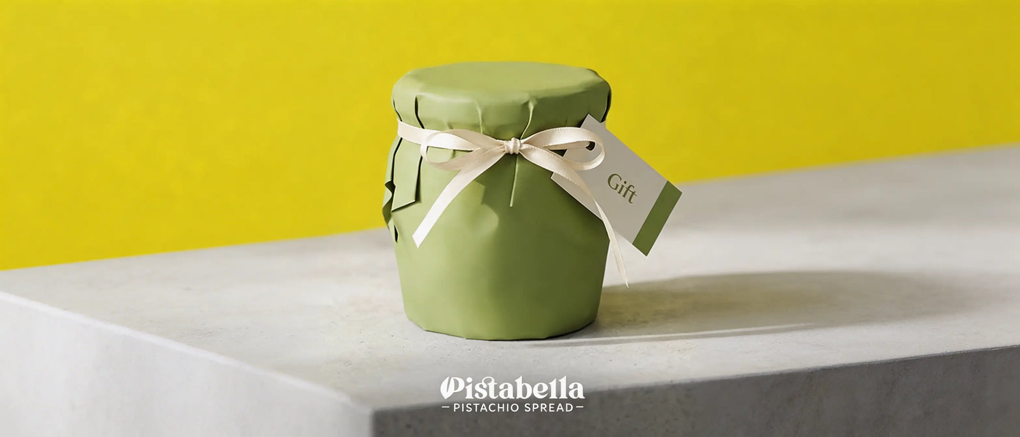 Pistabella jar wrapped in sage green paper with a cream ribbon and gift tag on a concrete shelf.